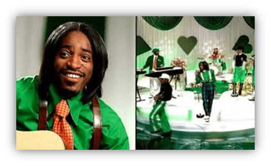 "Musician performing on stage in green attire with a band, featuring a vibrant set design."