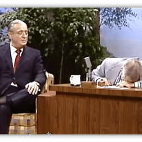 "Rodney Dangerfield performs comedy on a talk show, while the host laughs and leans on the desk."