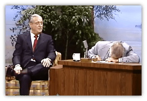 "Rodney Dangerfield performs comedy on a talk show, while the host laughs and leans on the desk."