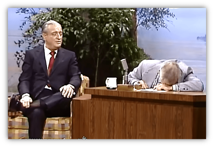 "Rodney Dangerfield performs comedy on a talk show, while the host laughs and leans on the desk."