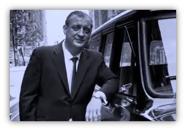 "Black and white photo of a man in a suit leaning against a classic car on a city street."