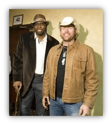 "Two men posing together, one in a dark blazer and the other in a cowboy hat and jacket, in a hallway."