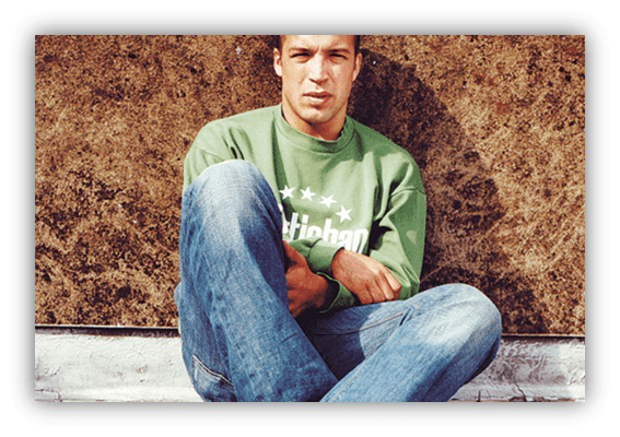 Young man sitting casually against a wall, wearing a green sweatshirt and jeans.