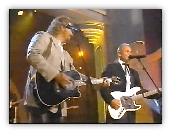 "Two musicians performing on stage, one with an acoustic guitar and the other with an electric guitar, under vibrant stage lights."