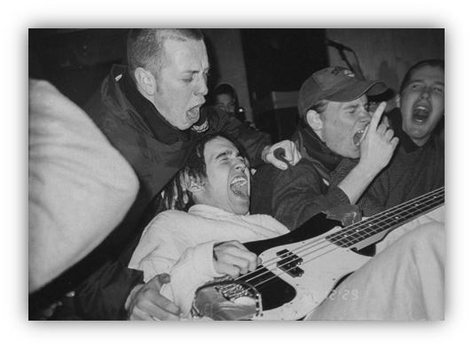 "Black and white image of a lively punk rock concert with fans passionately singing and playing bass."