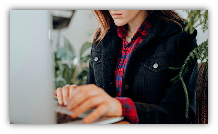 Woman typing on a laptop surrounded by plants, wearing a plaid shirt and denim jacket.