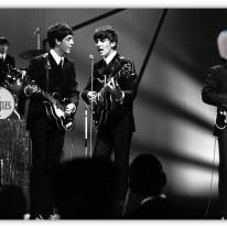 The Beatles performing live on stage in black and white.
