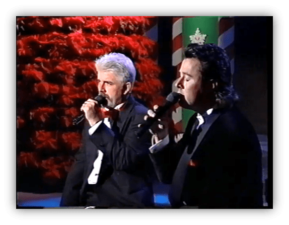 Two male singers in tuxedos performing on stage with a festive backdrop.