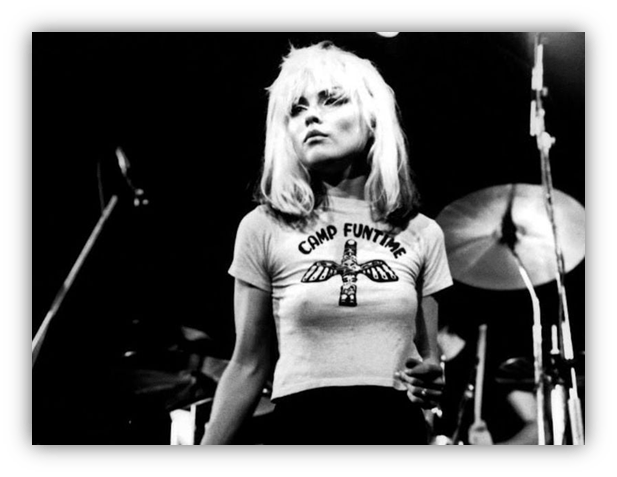 "Black and white photo of a female musician on stage wearing a 'Camp Funtime' shirt, with a drum set in the background."