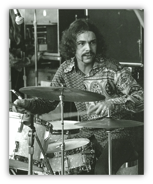 Black and white image of a drummer in a patterned shirt performing on stage.