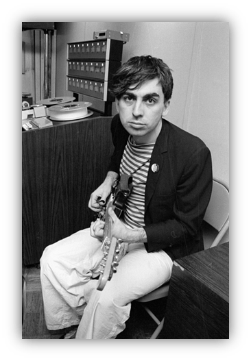 Musician sitting with guitar in a recording studio, wearing a striped shirt and blazer.