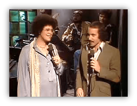 "Two singers performing on stage with microphones, accompanied by a band in the background."