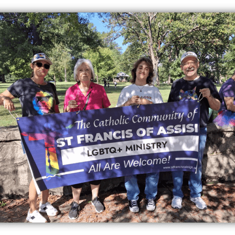 Four people holding a banner for St. Francis of Assisi LGBTQ+ Ministry, promoting inclusivity in a park setting.