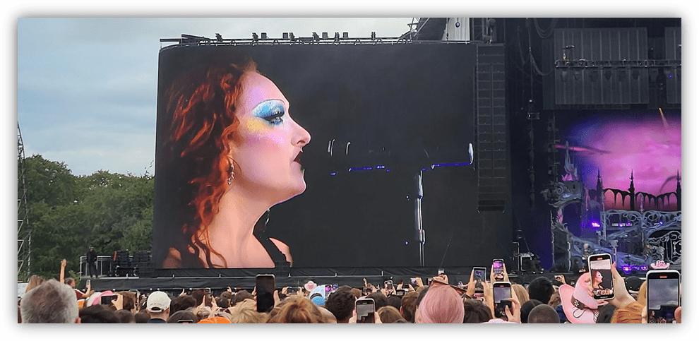 "Close-up of a singer with colorful makeup performing on stage, large screen in background, crowd capturing the moment."