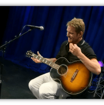 Musician performing with an acoustic guitar on stage.