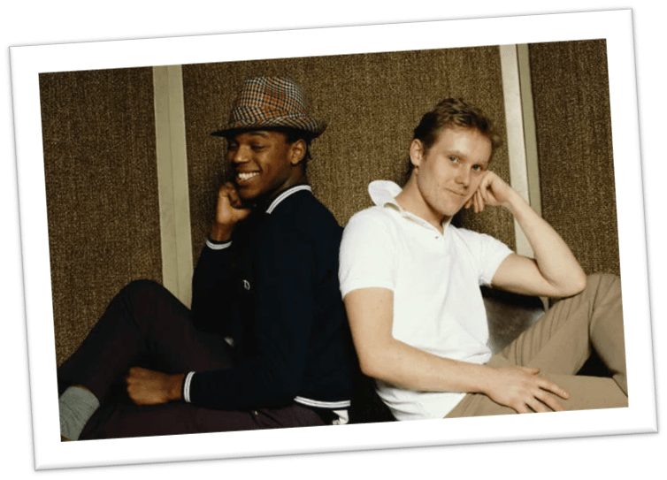 Two men seated back-to-back, one wearing a hat and stylish clothing, posing with smiles.
