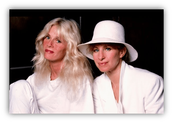 Two women in stylish white outfits, one with long blonde hair and the other wearing a white hat, pose together.