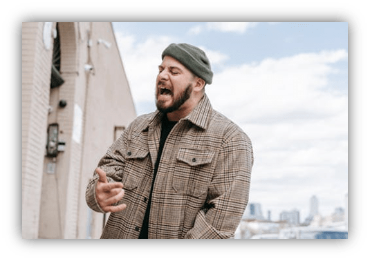 "Man passionately rapping outdoors in a plaid jacket and green beanie."