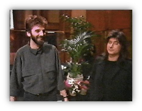 Two people smiling in a warmly lit indoor setting with plants and flowers.