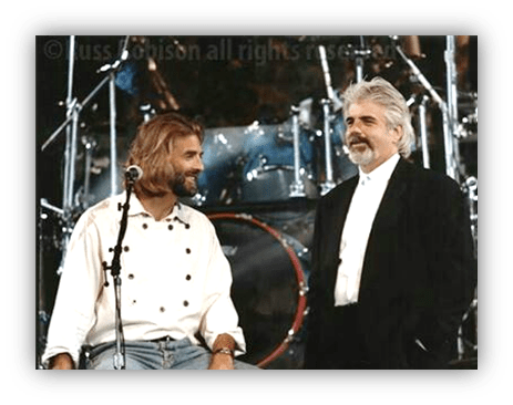 "Two musicians on stage, one in a white chef-style shirt and the other in a black suit, with drums in the background."