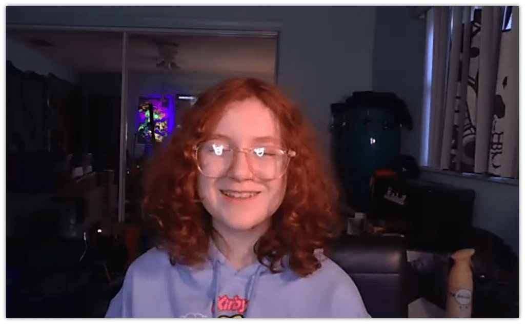 Smiling teenager with curly red hair wearing glasses, sitting in a cozy room with colorful lighting.
