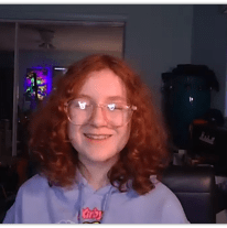 Smiling teenager with curly red hair wearing glasses, sitting in a cozy room with colorful lighting.