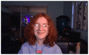 Smiling teenager with curly red hair wearing glasses, sitting in a cozy room with colorful lighting.