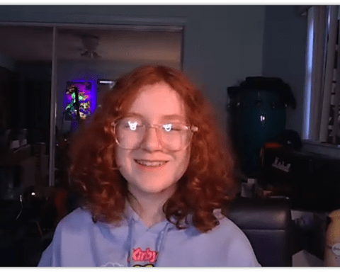 Smiling teenager with curly red hair wearing glasses, sitting in a cozy room with colorful lighting.
