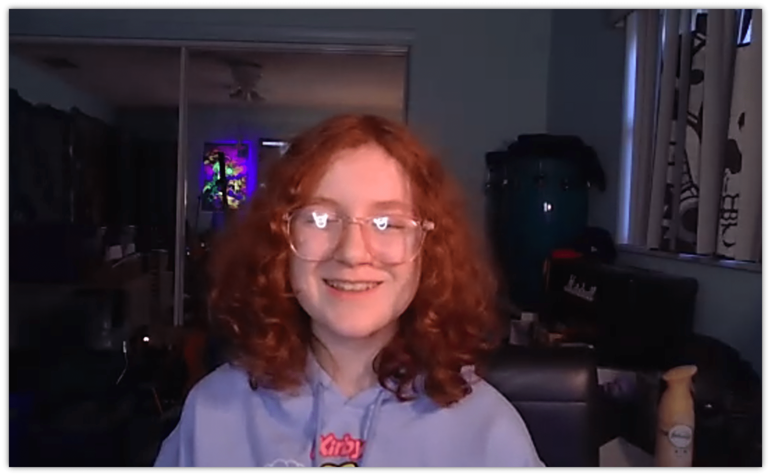 Smiling teenager with curly red hair wearing glasses, sitting in a cozy room with colorful lighting.