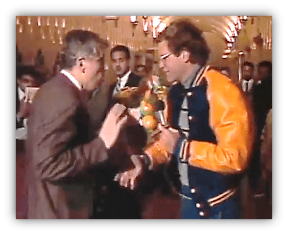 Two men engaged in a lively conversation at a formal event, with guests in the background.