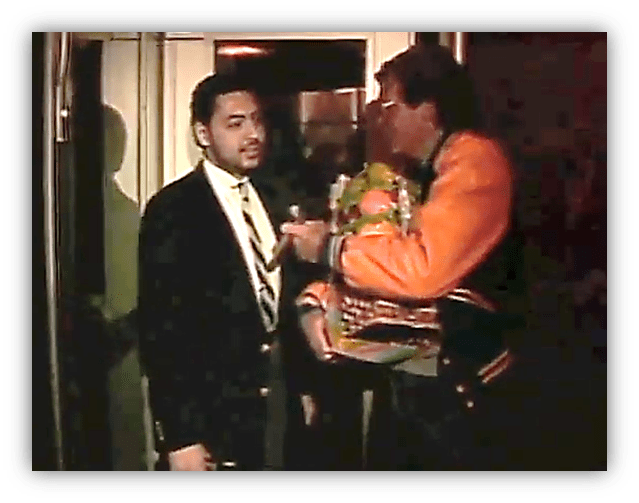 Two men at an event, one in a suit and the other in an orange jacket holding a bouquet.