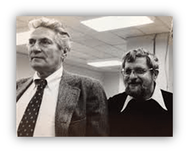 Black and white photo of two men, one in a suit and tie, the other in a sweater, standing in an office setting.