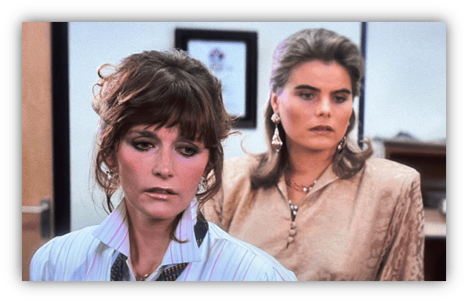 Two women in stylish 1980s attire, looking concerned in an office setting.