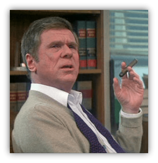 Man in a cardigan holding a cigar, looking thoughtfully at the camera with bookshelves in the background.