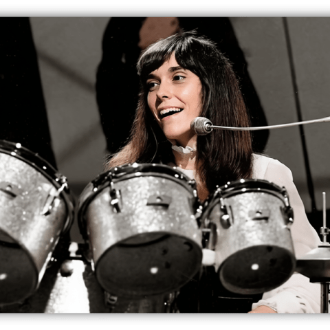 "Smiling female drummer performing with a silver drum set on stage."