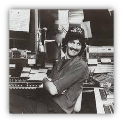 Radio DJ in a studio with equipment, smiling and wearing a hat.