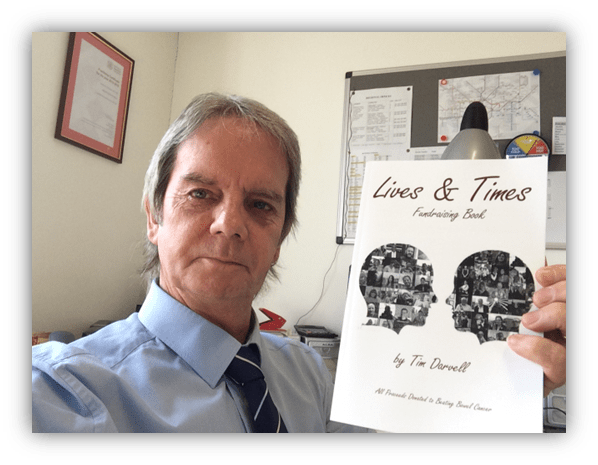 Man holding "Lives & Times" fundraising book by Tim Darwell in an office setting.