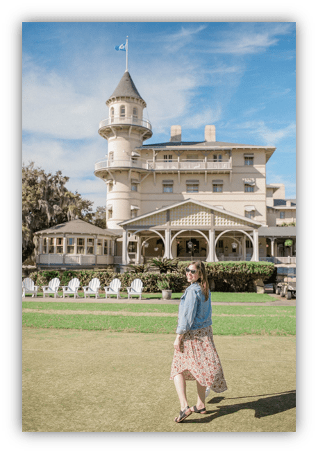 "Woman in floral dress and denim jacket walking in front of historic hotel with tower."