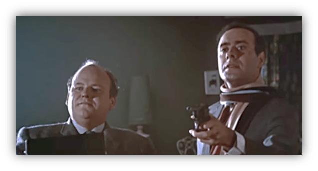 Two men in a tense scene, one holding a gun while the other looks on, set in a dimly lit room.