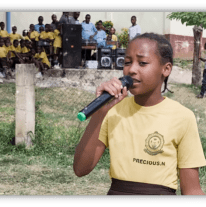 Young girl singing with a microphone at a school event, surrounded by students in yellow uniforms.