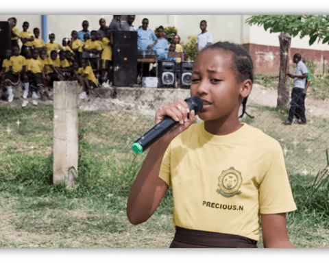 Young girl singing with a microphone at a school event, surrounded by students in yellow uniforms.