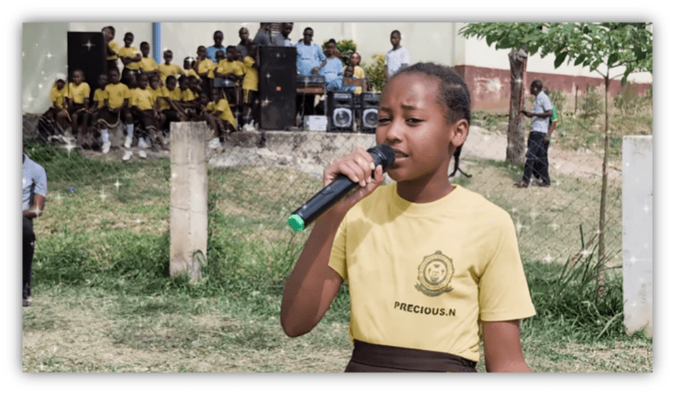 Young girl singing with a microphone at a school event, surrounded by students in yellow uniforms.
