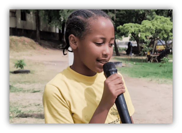 Young girl in a yellow shirt speaking into a microphone outdoors.