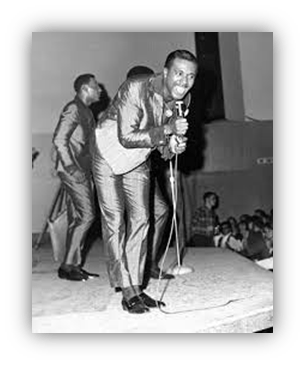 "Black and white photo of a singer performing on stage in a shiny suit, with a microphone in hand and a lively audience in the background."