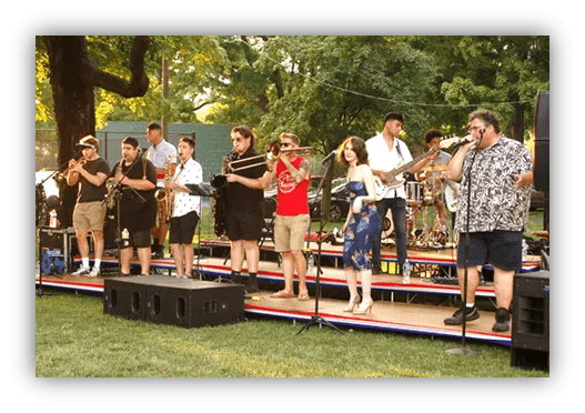 Live music performance featuring a diverse band with brass instruments, vocals, and a lively audience in a park setting.