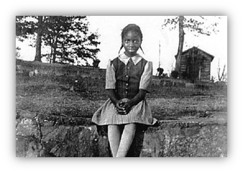 "Historic black and white photo of a young girl in a dress sitting outdoors."