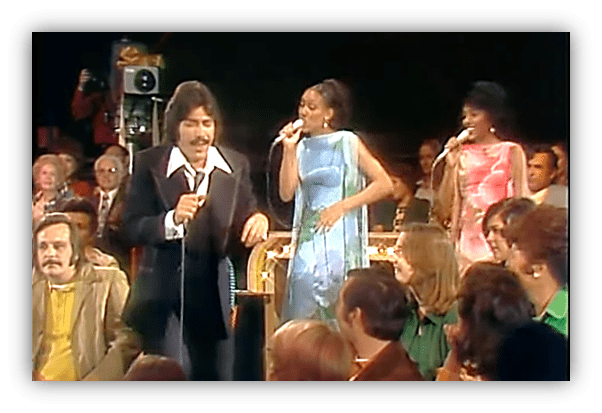"Male singer performing on stage with two female backup singers in colorful dresses, audience enjoying the show."