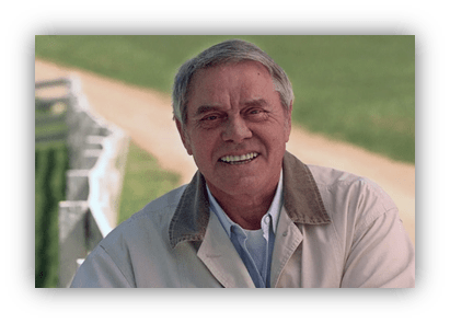 Smiling older man in a light jacket outdoors, with a green field and path in the background.