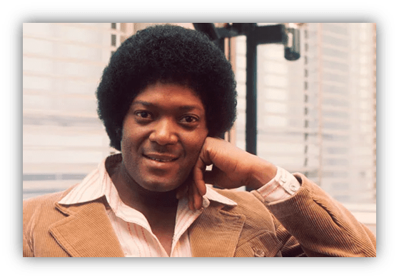 "Smiling man with an afro hairstyle, wearing a brown corduroy jacket and a white shirt, seated indoors."