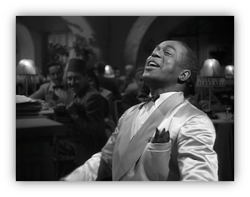 "Man in a white suit singing at a vintage café, with an audience in the background."